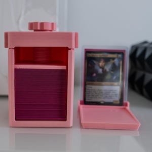 May include: A pink plastic card storage box with a clear lid and a compartment for holding cards. The box is open and a stack of cards is visible inside.