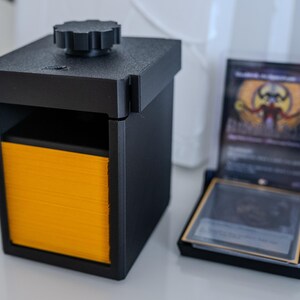 May include: A black 3D printed card storage box with a yellow interior and a knob on the top. The box is designed to hold a deck of trading cards.