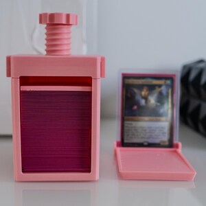 May include: A pink plastic card storage box with a screw-on top and a separate pink plastic card holder. The card holder has a clear window to view the cards inside.