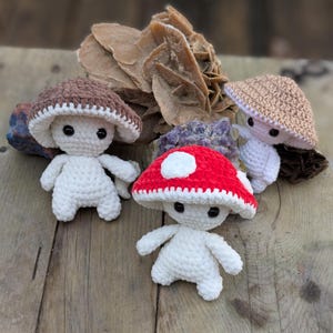 Puede incluir: Tres figuras de hongos de ganchillo hechas a mano con cuerpos blancos y sombreros de diferentes colores: marrón, rojo con lunares blancos y beige. Los hongos se exhiben sobre una superficie de madera con formaciones rocosas naturales en el fondo. Las figuras tienen ojos negros.