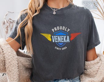 May include: Dark gray t-shirt with the text "PROUDLY VENEKA SINCE 2004" in a shield design. The shield has yellow and red wings. The shirt is a pepper color.