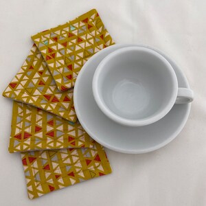 May include: Four yellow, red, and blue geometric patterned fabric coasters with a white ceramic teacup and saucer on top.