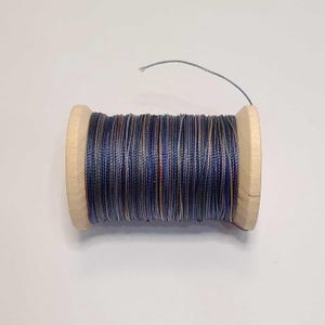 May include: A wooden spool wrapped with multi-colored thread featuring a blend of dark blue, grey, and subtle orange tones. The thread is wound tightly around the cylindrical wooden core, with a single loose strand extending from the top edge.