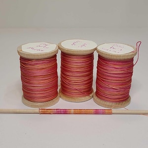 May include: Three wooden spools wrapped in variegated thread featuring shades of pink, orange, and red. Each spool has a white paper label on top with handwritten text. A single wooden dowel is positioned horizontally in front of the spools.