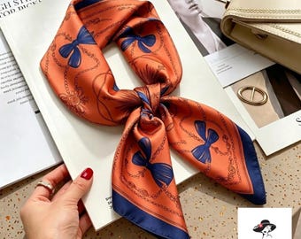 May include: A square scarf featuring an orange background with a dark blue border and a repeating pattern of dark blue bows and thin chain links. The scarf is tied in a knot, displaying the vibrant orange fabric and the contrasting blue details.
