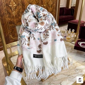 May include: A white scarf featuring an intricate embroidered floral pattern in shades of pink, blue, and brown. The scarf has long, soft fringe detailing at the bottom edge and a small black rectangular label with a goat icon and text.
