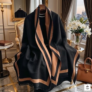 Op de afbeelding: Een zwarte sjaal met een brede beige rand en franjes, gedrapeerd over een donker kledingstuk. De sjaal heeft een effen zwart middenpaneel met een contrasterende beige streep langs de randen en de zoom, wat zorgt voor een gestructureerde uitstraling.