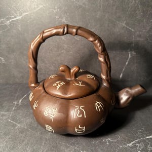 Puede incluir: Tetera de cerámica marrón con asa y boquilla de estilo bambú. La tetera tiene un cuerpo redondeado con tapa y está decorada con caracteres chinos blancos. La tetera está diseñada para preparar té.