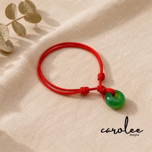 May include: A bracelet featuring a vibrant red cord with adjustable knots and a smooth, circular green jade-like stone pendant. The design includes a central ring-shaped charm suspended from the red cord, with the text carolee designs visible.