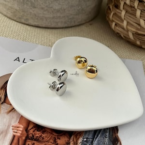 May include: A white heart-shaped dish holds two pairs of earrings. One pair is silver, oval-shaped studs, and the other is gold, round studs. The word "cardee" is written in cursive on the dish. The word "ALT" is partially visible on a magazine.