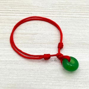 May include: A red string bracelet with a green jade bead. The bead is round and has a hole in the center. The bracelet is tied with a knot.