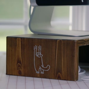May include: A wooden computer monitor stand with a cat graphic. The stand is brown and has a cutout for cords. A laptop is sitting on the stand.