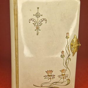 May include: A white book with a gold trim and floral designs. The book has a gold clasp with a decorative knob.