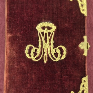 May include: A rectangular book bound in deep burgundy velvet featuring ornate gold-toned metal corner protectors and a central gold-toned metal monogram emblem. The cover includes a small gold-toned metal clasp on the right side and metal studs.