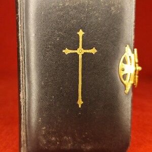 May include: A small, black book with a gold cross emblem and a gold clasp. The book has a red tassel and appears to be made of leather. The cover is slightly worn, suggesting age.