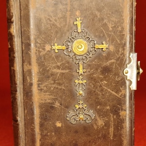 Könnte beinhalten: Ein Vintage-Buch in braunem Leder mit einem dekorativen Kreuzdesign. Das Kreuz besteht aus gold- und silberfarbenem Metall mit aufwendigen Details. Ein Metallverschluss sichert den Verschluss des Buches.