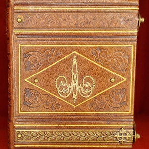May include: A vintage, leather-bound book with intricate gold detailing. The cover features a diamond-shaped emblem with ornate gold filigree and is framed by gold borders. The book's edges are adorned with decorative patterns and gold accents.
