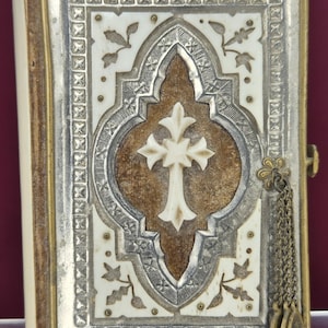 May include: An antique book with a decorative cover. The cover features a white cross on a brown background, surrounded by ornate silver and ivory-colored details. A small chain with three medallions hangs from the side.