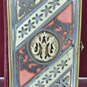 Può includere: Una copertina di libro rettangolare con un medaglione circolare centrale e un emblema intagliato color crema. La copertina è decorata con fasce diagonali color crema, filigrana in metallo scuro e pannelli color salmone.