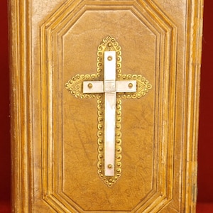 May include: An antique book cover in gold and tan tones, with a decorative cross as the central feature. The cross is white with gold detailing. The cover has a textured finish and a geometric border design.