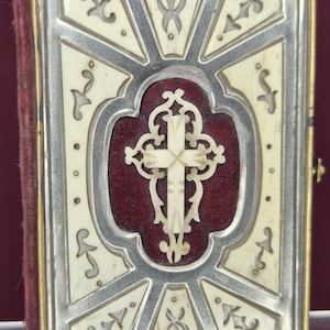 May include: A small, rectangular religious book with a cream-colored cover and a silver frame. The cover features an ornate cross design and decorative patterns. The book's spine is a deep red color, and the edges are trimmed with gold.