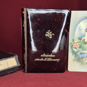 May include: A collection of vintage religious items, including a dark brown book with gold lettering and a decorative emblem, a small box with a label, and two religious cards with illustrations.
