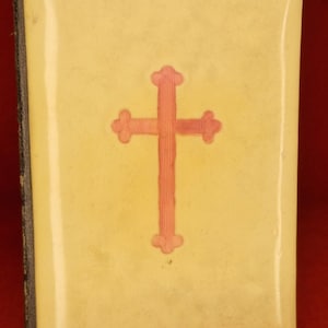 May include: A small, rectangular book with a pale yellow cover and a pink cross design. The book's spine is dark, and the cover has a glossy finish. The cross is centered on the cover.