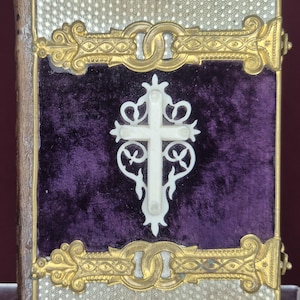 May include: A small, antique book with a silver cover and gold filigree accents. The center features a deep purple velvet panel with a white cross. The book's edges are gold, and it has a clasp.