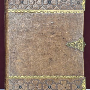 Puede incluir: Un libro encuadernado en cuero marrón con patrones de trébol en relieve en los bordes superior e inferior. La cubierta presenta esquineros de metal dorado y un cierre metálico central en el lado derecho, con una superficie texturizada.