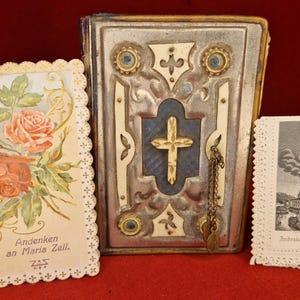 Puede incluir: Un libro antiguo con cubierta plateada, cruz dorada y cadena, flanqueado por dos tarjetas vintage. Una tarjeta presenta rosas y el texto "Andenken an Maria Zell." La otra tarjeta muestra una iglesia.