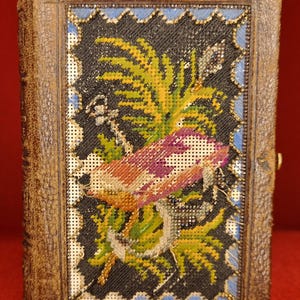 Puede incluir: Libro antiguo encuadernado en cuero con una cubierta de punto de aguja detallada. La cubierta presenta un diseño floral con tonos verdes, amarillos y morados sobre un fondo negro, enmarcado por un borde azul. El libro tiene un cierre de latón.