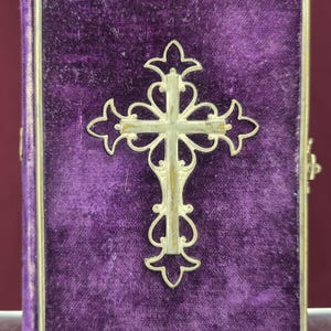 May include: A small, rectangular book with a deep purple velvet cover. A silver-colored, ornate cross is centered on the cover. The book has a gold-colored metal frame and a clasp on the right side.