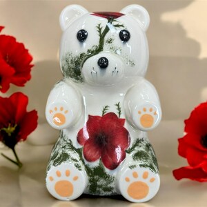 May include: A white ceramic bear figurine with a red poppy design on its chest and head. The bear has black eyes, orange paw pads, and green accents. Red poppies are in the background.