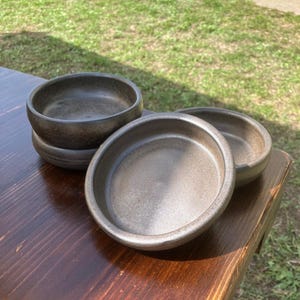 Peut inclure: Quatre petits bols ronds en céramique gris-brun. Les bols ont un design simple et minimaliste avec une surface légèrement texturée. Ils sont disposés sur une surface en bois foncé, avec de l'herbe en arrière-plan.