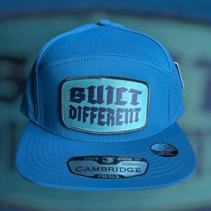 Puede incluir: Una gorra de béisbol azul cerceta con un parche rectangular en la parte delantera con el texto negro BUILT DIFFERENT. La gorra tiene paneles laterales perforados, una visera plana y una pegatina en la visera que dice SINCE 2000 CAMBRIDGE PB313 HYBRID.