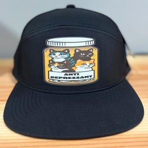 May include: Black baseball cap featuring a graphic of four cats inside a jar with the words "ANTI DEPRESSANT". The cats are various colors, including black, orange, and blue. The cap has a curved brim and a button on top.