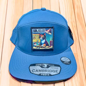 May include: Blue hybrid baseball cap with a flat brim. The cap features a square patch with a cat illustration and the text "ADL MEOW & MORE CREATIONS". The cap also has the text "CAMBRIDGE" and "PB313".