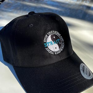 May include: Black baseball cap with the text "STOP THE RACISM AND IGNORANCE TODAY" embroidered on the front. The design includes a yin-yang symbol and the word "STRAIT" in teal.