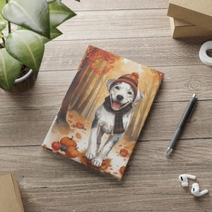 Könnte beinhalten: Ein Notizbuch mit einer Aquarellillustration eines Hundes mit Hut und Schal in einer Herbstwaldszene. Kürbisse und Blätter sind um den Hund herum verstreut. Das Wort "Pup" steht oben auf dem Notizbuch.