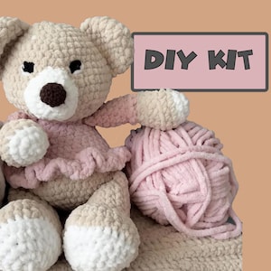 May include: A crocheted teddy bear made of beige and white plush yarn, wearing a pink ruffled top, positioned next to a ball of light pink yarn. A rectangular sign with a pink background and black text reads DIY KIT in the upper right corner.