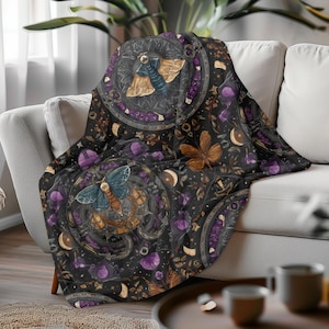 May include: A black throw blanket with a detailed pattern featuring moths, moons, and floral elements in shades of blue, brown, purple, and gold. The blanket is draped over a white sofa.