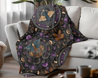 May include: A black throw blanket with a detailed pattern featuring moths, moons, and floral elements in shades of blue, brown, purple, and gold. The blanket is draped over a white sofa.