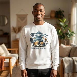 Puede incluir: Sudadera blanca con un gráfico de un perro durmiendo bajo una nube, rodeado de iconos tecnológicos. El texto "SYSTEM FAILURE, NAP SUCCESS" está debajo. La persona lleva pantalones negros.