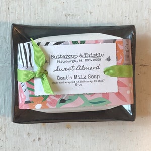 Puede incluir: Una pastilla de jabón de leche de cabra envuelta en papel estampado con flores rosas y verdes con una cinta verde. El jabón está etiquetado con "Buttercup & Thistle", "Sweet Almond", "Goat's Milk Soap" y "Made and wrapped in McMurray, PA 15317".