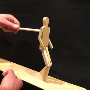May include: A wooden toy figure with articulated legs and arms, standing on a wooden board. The figure is being moved by a hand holding a wooden stick.