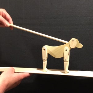 May include: A wooden toy dog with jointed legs is being walked on a wooden platform by a hand holding a stick. The dog is light brown and has a white circle for an ear.