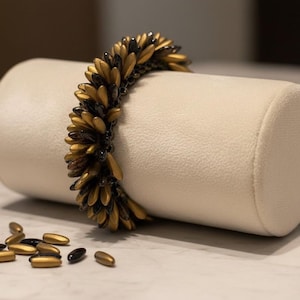 May include: A gold and black beaded bracelet is displayed on a cream-colored cylindrical jewelry holder. The bracelet features a repeating pattern of gold and black beads. Several loose beads are scattered nearby.