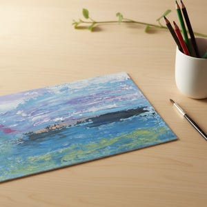 May include: An abstract painting with blue, purple, and green brushstrokes. A white ceramic cup holds colored pencils. A paintbrush with a black handle rests nearby on a light wooden surface. A sprig of greenery is in the background.