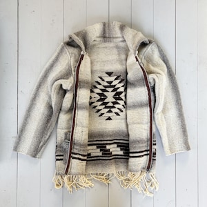 Puede incluir: Chaqueta con capucha gris y blanca con un diseño geométrico en el centro. La chaqueta tiene cierre de cremallera y flecos en la parte inferior. La chaqueta está hecha de un material tejido.