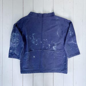 May include: A navy blue work jacket with short sleeves and a large pocket. The jacket is splattered with white paint. The jacket is laid flat on a white wooden surface.
